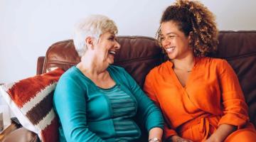 An older woman sits with a younger woman, laughing on a sofa
