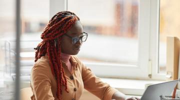 A woman wearing glasses sits at a laptop next to a large window