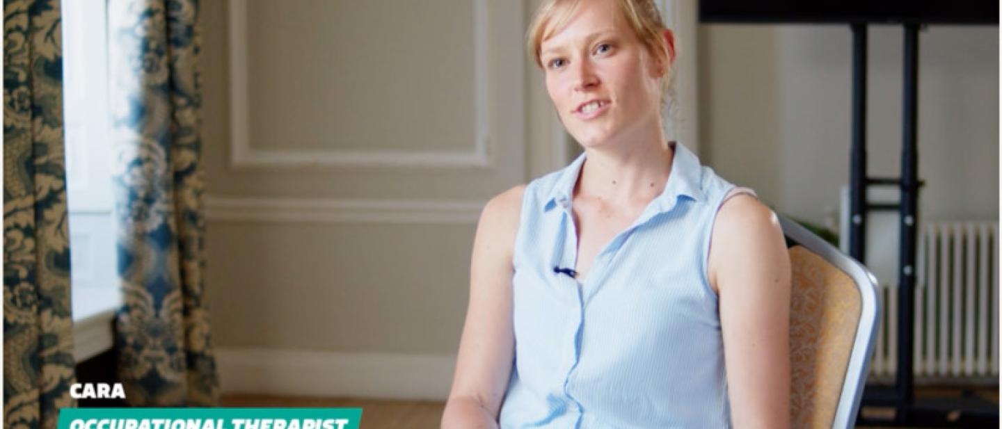 A lady in a blue vest top sat on a chair.  Her name is Cara