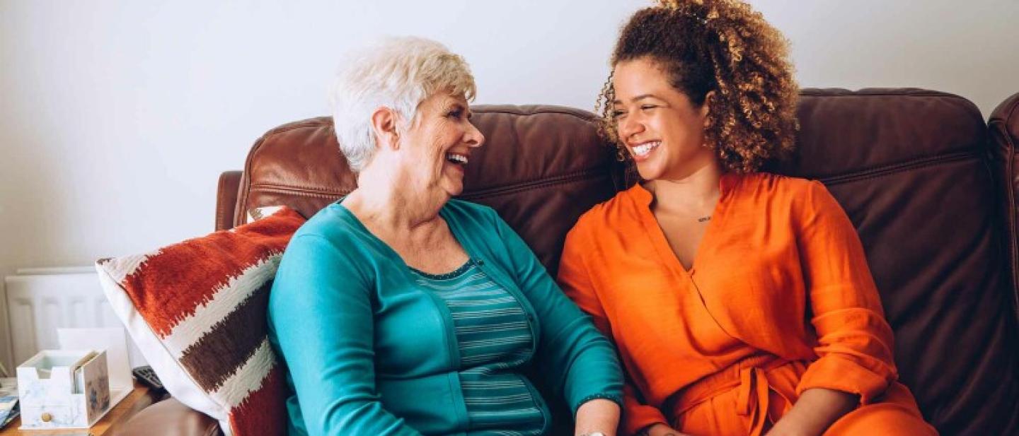 An older woman sits with a younger woman, laughing on a sofa