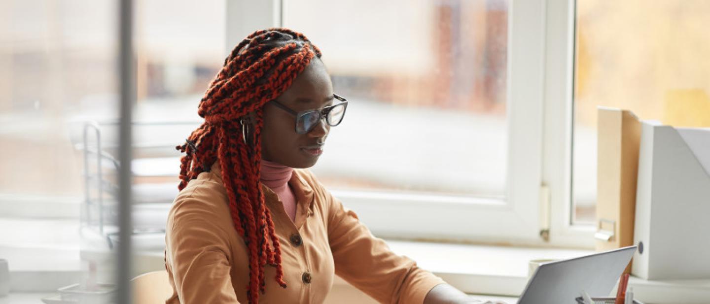 A woman wearing glasses sits at a laptop next to a large window